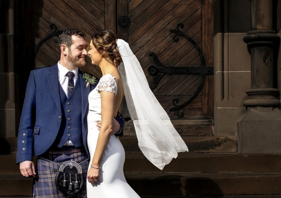 Bride wearing veil and groom wearing a kilt standing outside venue in an embrace while smiling at each other